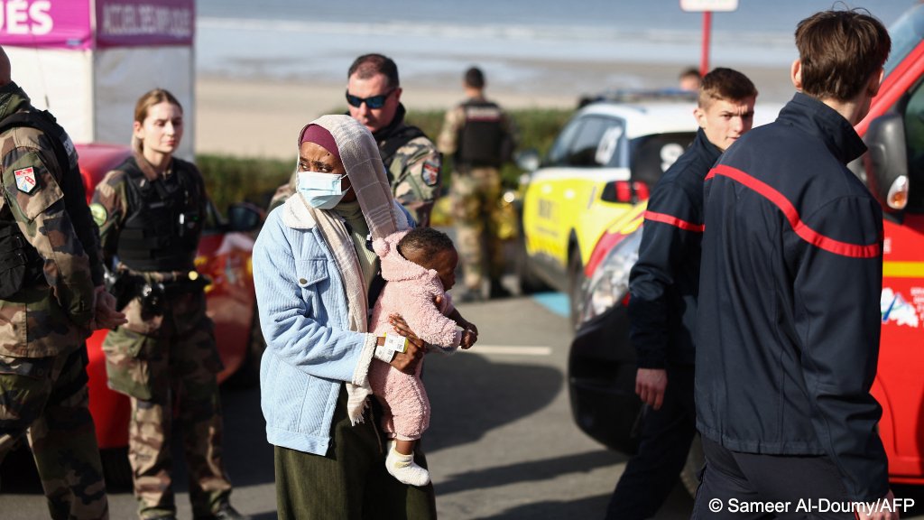 A woman holding a baby is being looked after by the emergency services after the deaths of four migrants while trying to board a 'taxi boat' off Équihen plage on Thursday morning, 37 were brought ashore, 30 more reportedly tried to continue towards the UK | Photo: Sameer Al-Doumy / AFP
