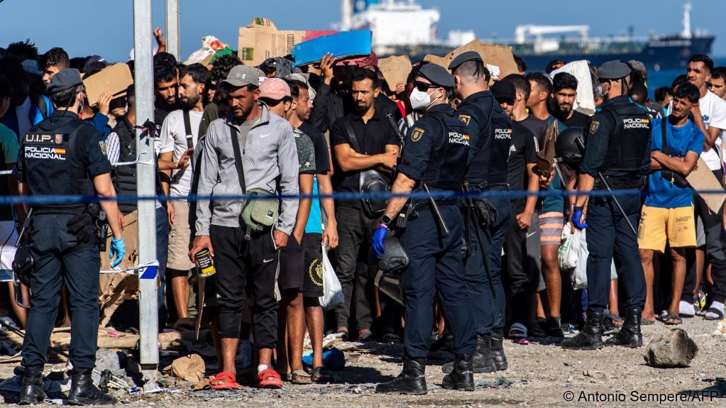 Police officers stand by as migrants wait for a food distribution in a temporary makeshift camp on an esplanade near the Trampolin beach while waiting to be transferred to reception facilities in Spain's north African enclave of Ceuta on October 7, 2026 | Photo: Antonio Sempere/AFP