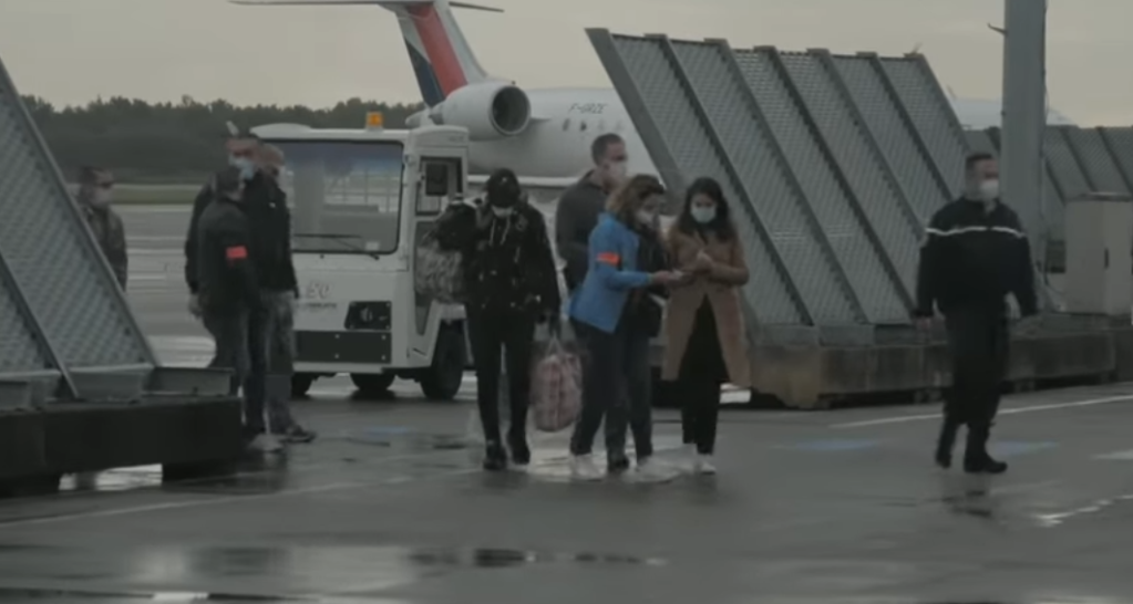 Ismail in the center, holding his bags is accompanied by more than 15 officials and police as he exits a deportation flight from the UK to France on which he was the only passenger | Source: Screenshot Channel 4 News