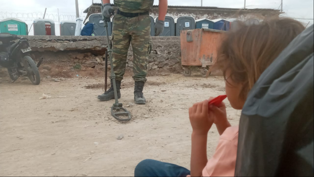 A Syrian girl looks on whilst a member of the Greek military appears to be searching for munitions after migrants have moved into the camp | Source: HRW / Private