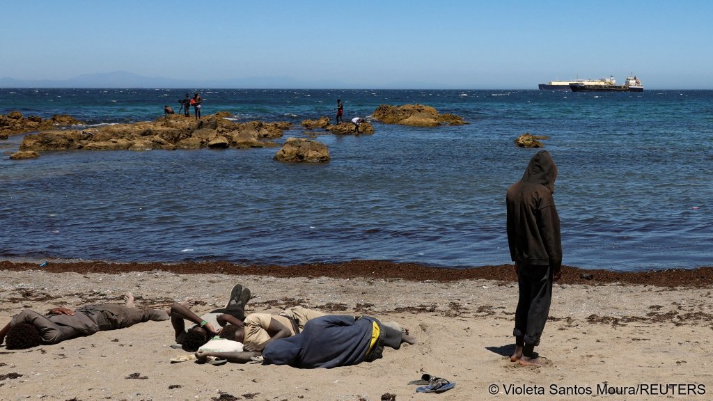 A migrant prays and another rests while waiting on the beach following mass crossings of migrants on foot and by sea from Morocco into the Spanish territory | Photo: Violeta Santos Moura / Reuters