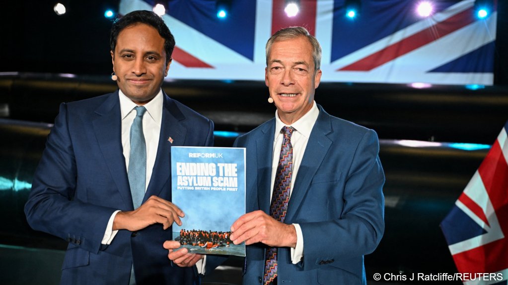 Reform UK's spokesperson for Home Affairs Zia Yusuf (l) and Reform UK leader Nigel Farage (r) hold a document titled 'ending the asylum scam, putting British people first' at a press conference, with a model of a dinghy visible in the background, in Hythe, Kent, Britain, October 12 | Photo: Chris J Ratcliffe / Reuters