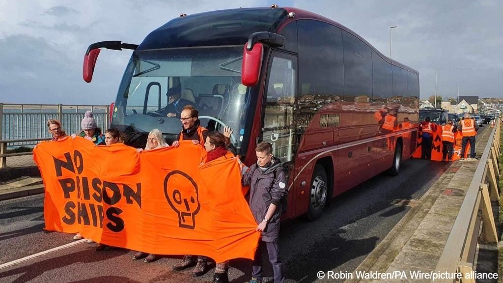 Just Stop Oil protesters block the bus carrying asylum seekers to board the Bibby Stockholm on Thursday (October 19), their banner reads 'No Prison Ships' | Photo: Robin Waldren / PA Wire / picture alliance